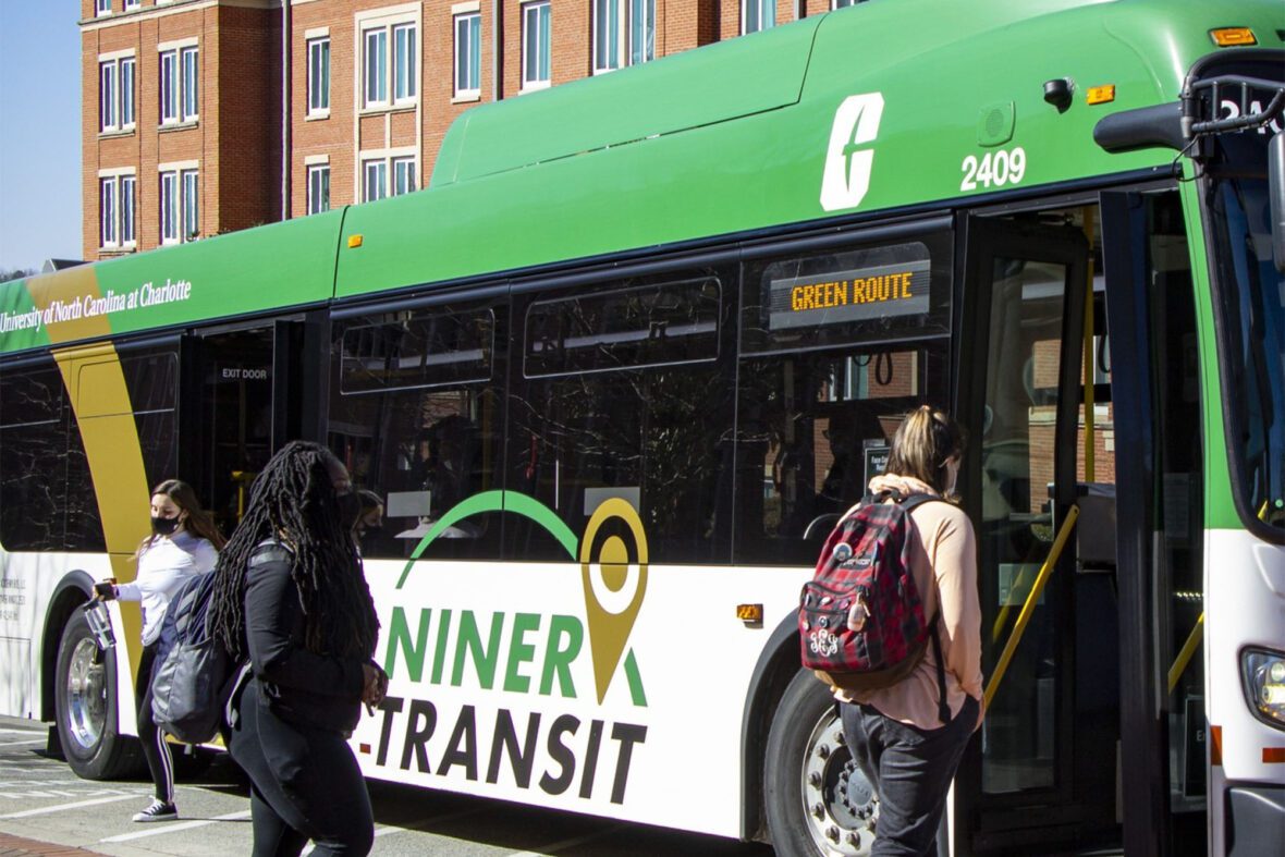 People boarding a Niner Transit bus on the Green Route in front of a brick building.