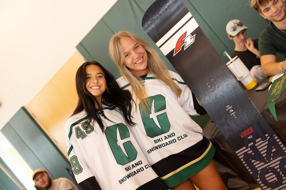Two people in matching jerseys pose with a snowboard at an indoor event.
