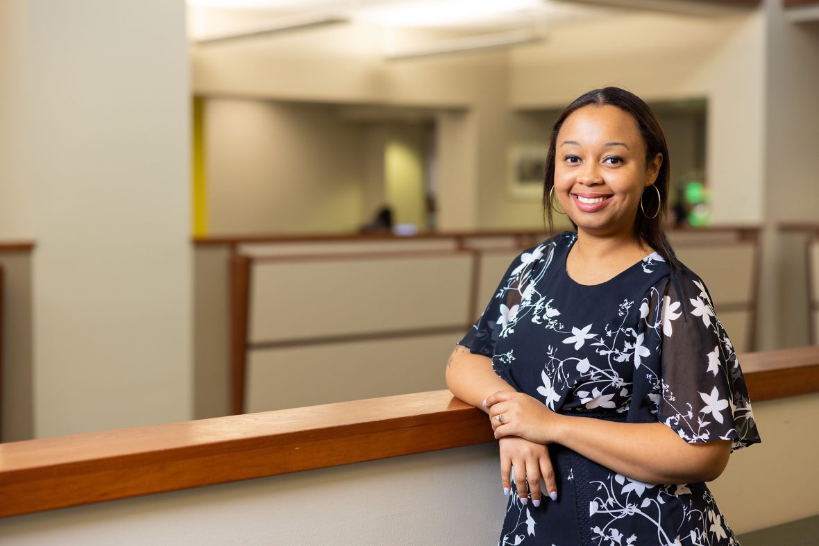 A woman in a black-and-white floral dress smiles while leaning on a railing inside an office building with an open atrium and cubicles in the background.