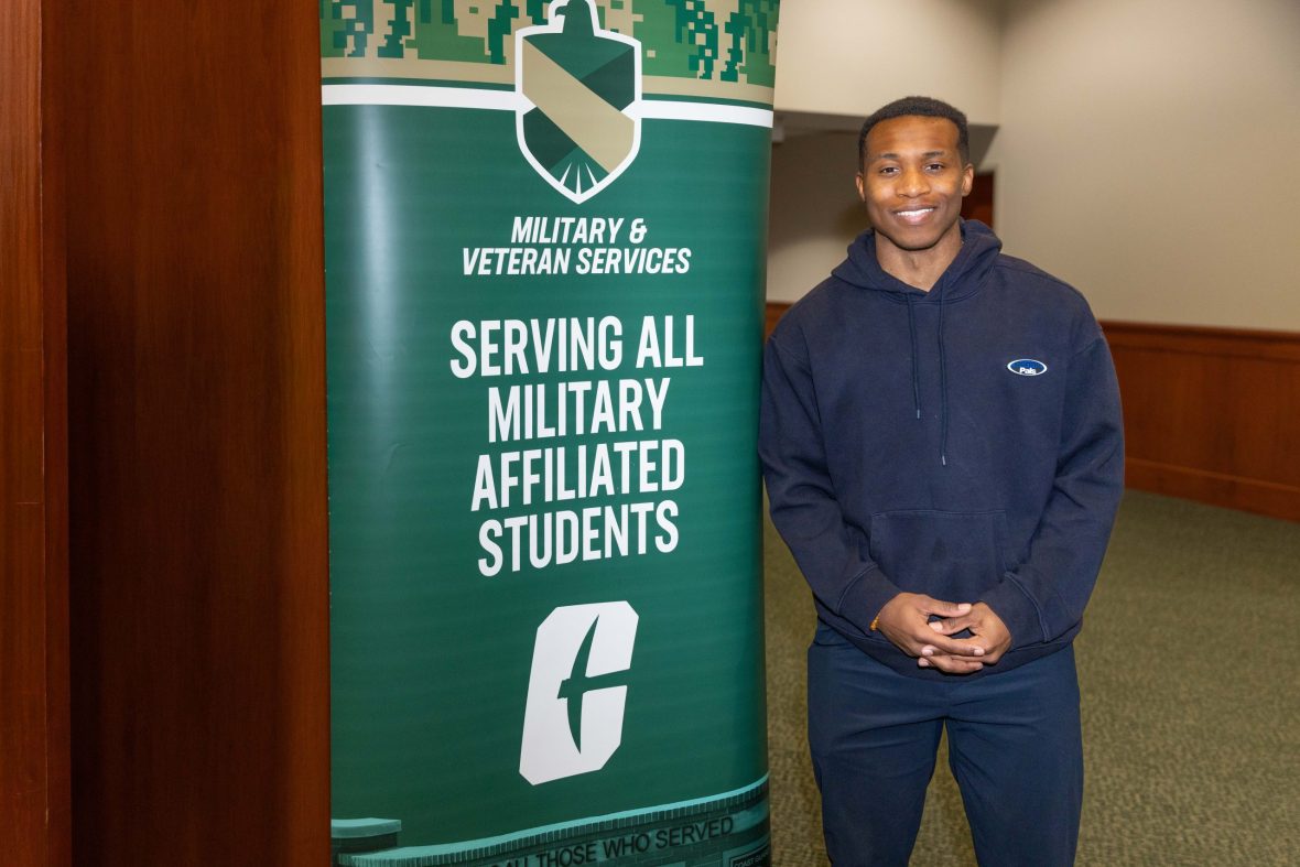 Smiling student stands beside a green Military & Veteran Services banner reading “Serving All Military Affiliated Students.”