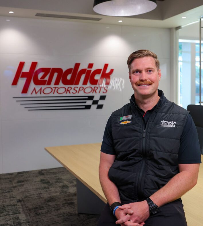 Smiling man wearing a black Hendrick Motorsports vest sits in a conference room with the red Hendrick Motorsports logo displayed on the wall behind him.